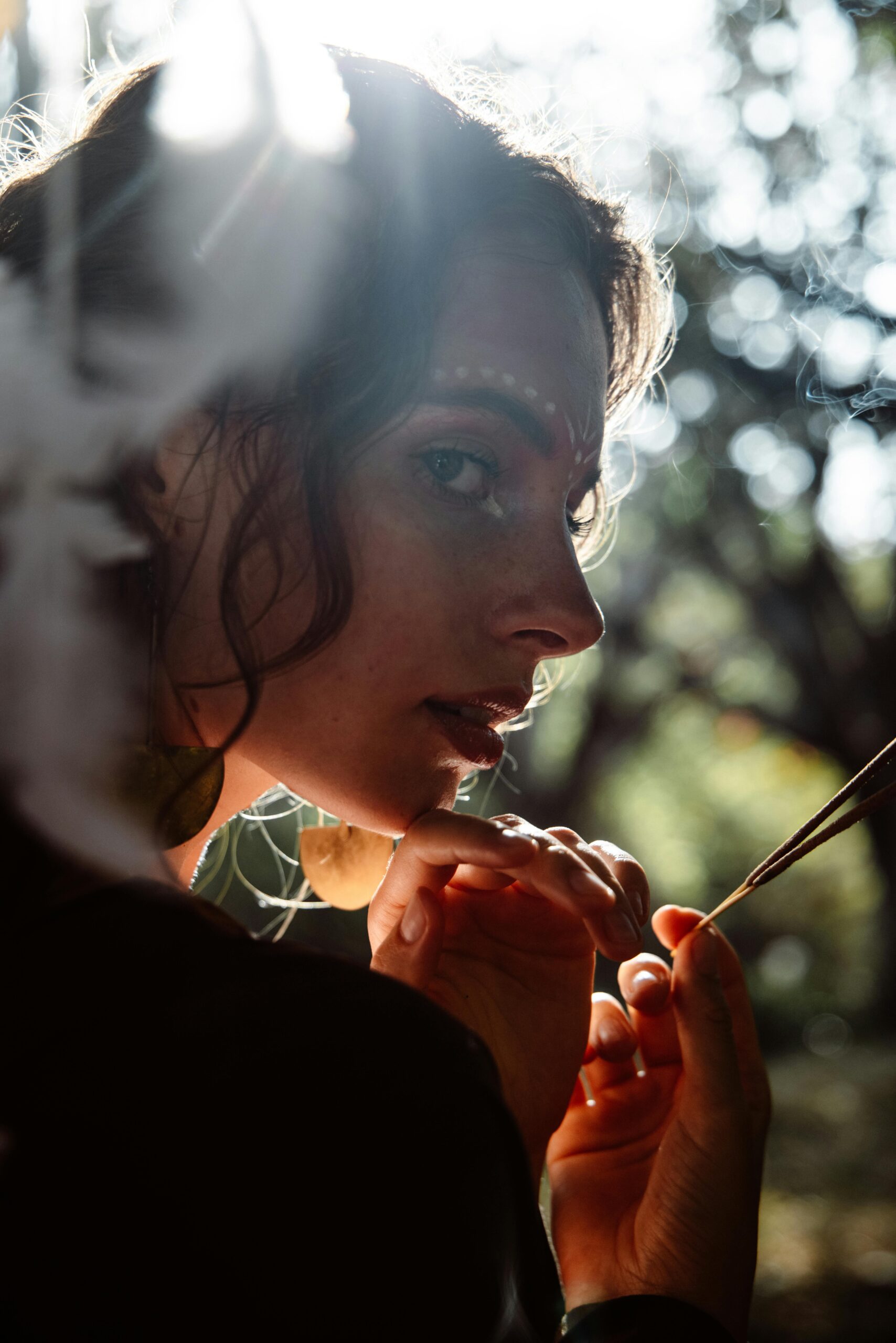 Mystical scene of a woman with makeup holding incense outdoors, in soft ethereal light.