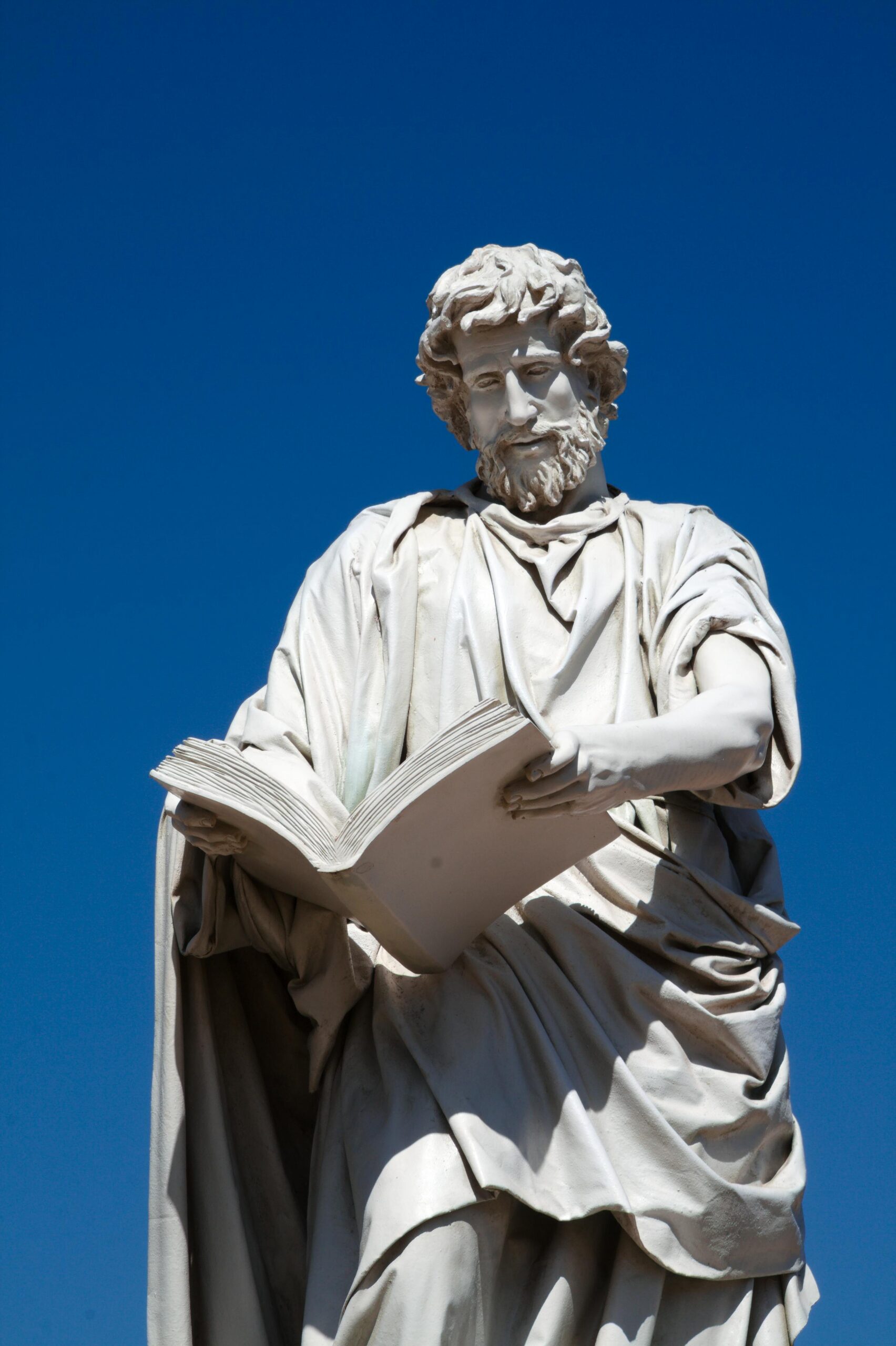 A marble statue of an apostle reading a book against a clear blue sky.