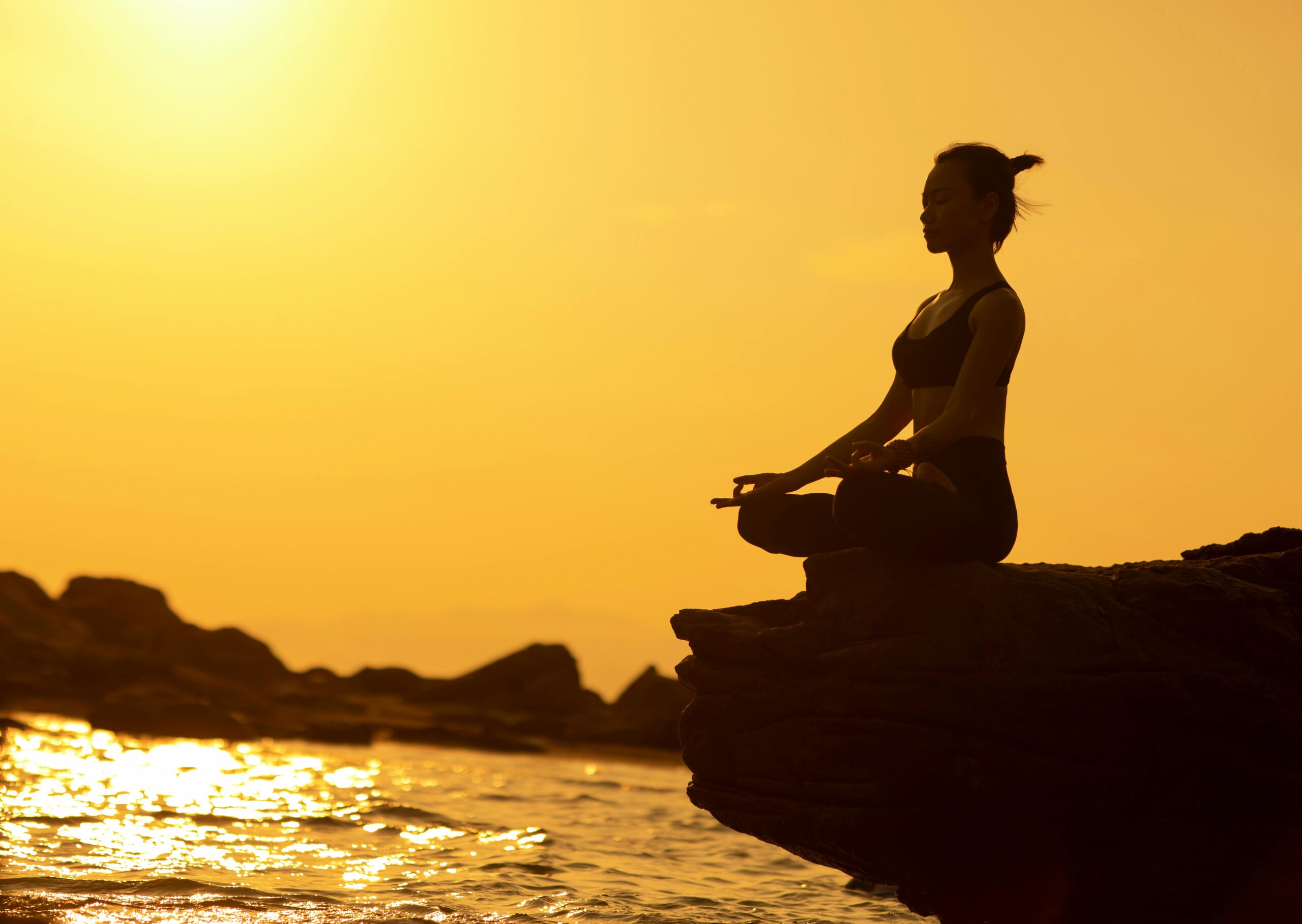 Woman practicing yoga in lotus pose on a cliff by the sea at sunset, embodying tranquility and balance.