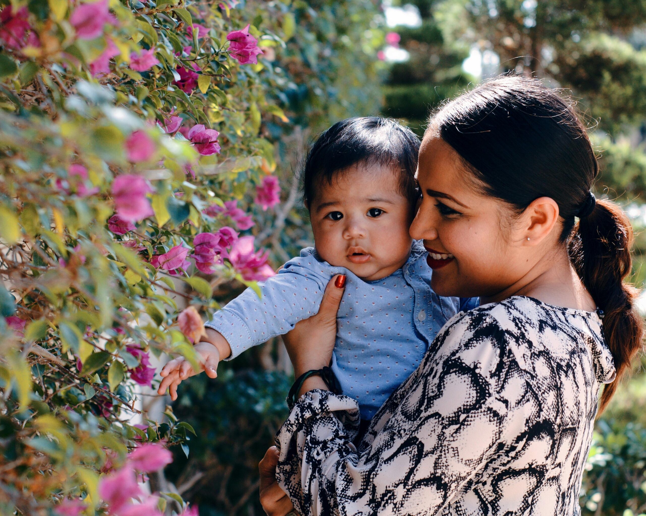 A smiling mother holds her baby near vibrant pink flowers in a sunny garden.