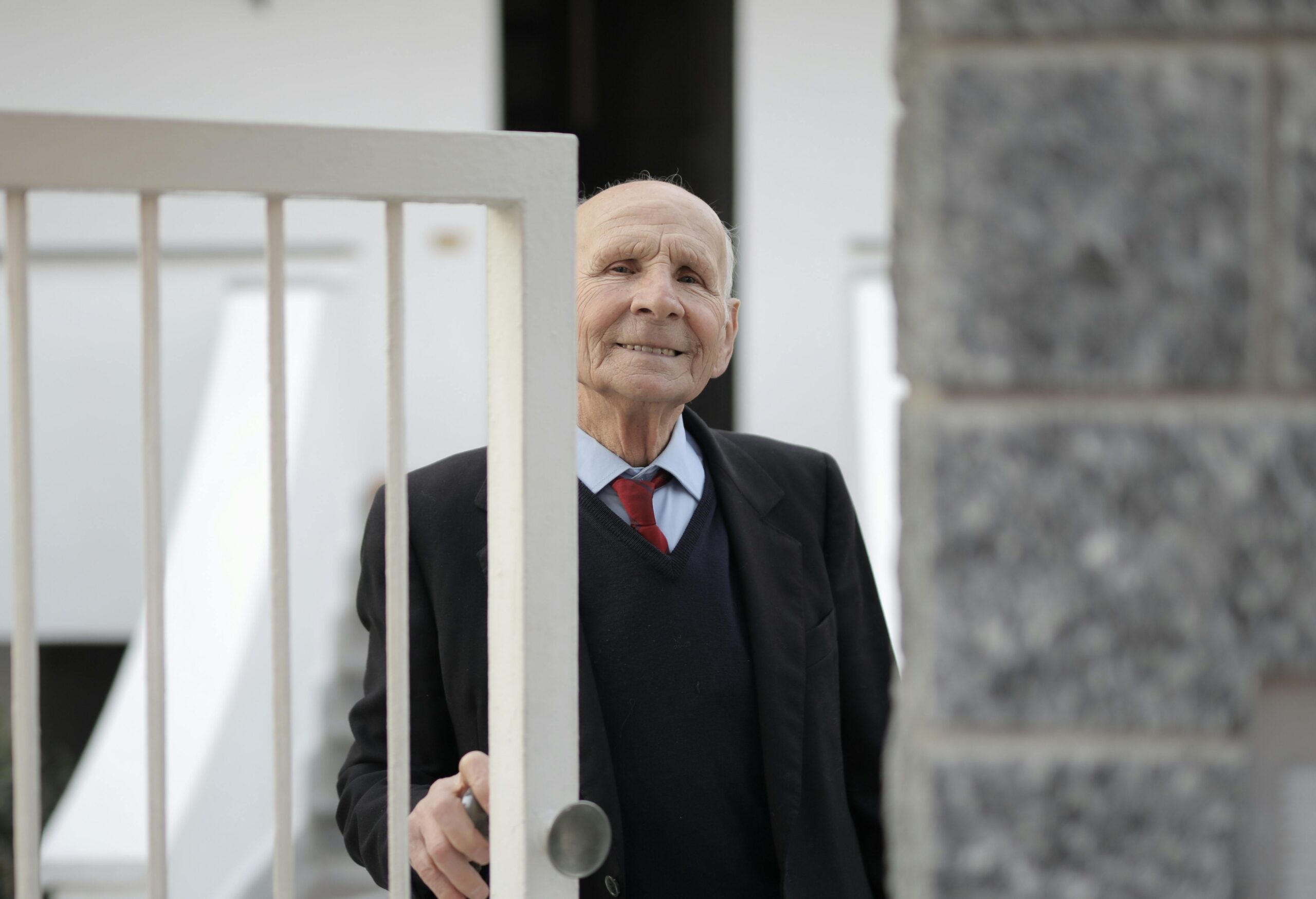 Cheerful elderly man in a suit smiling beside an open gate, outdoors.
