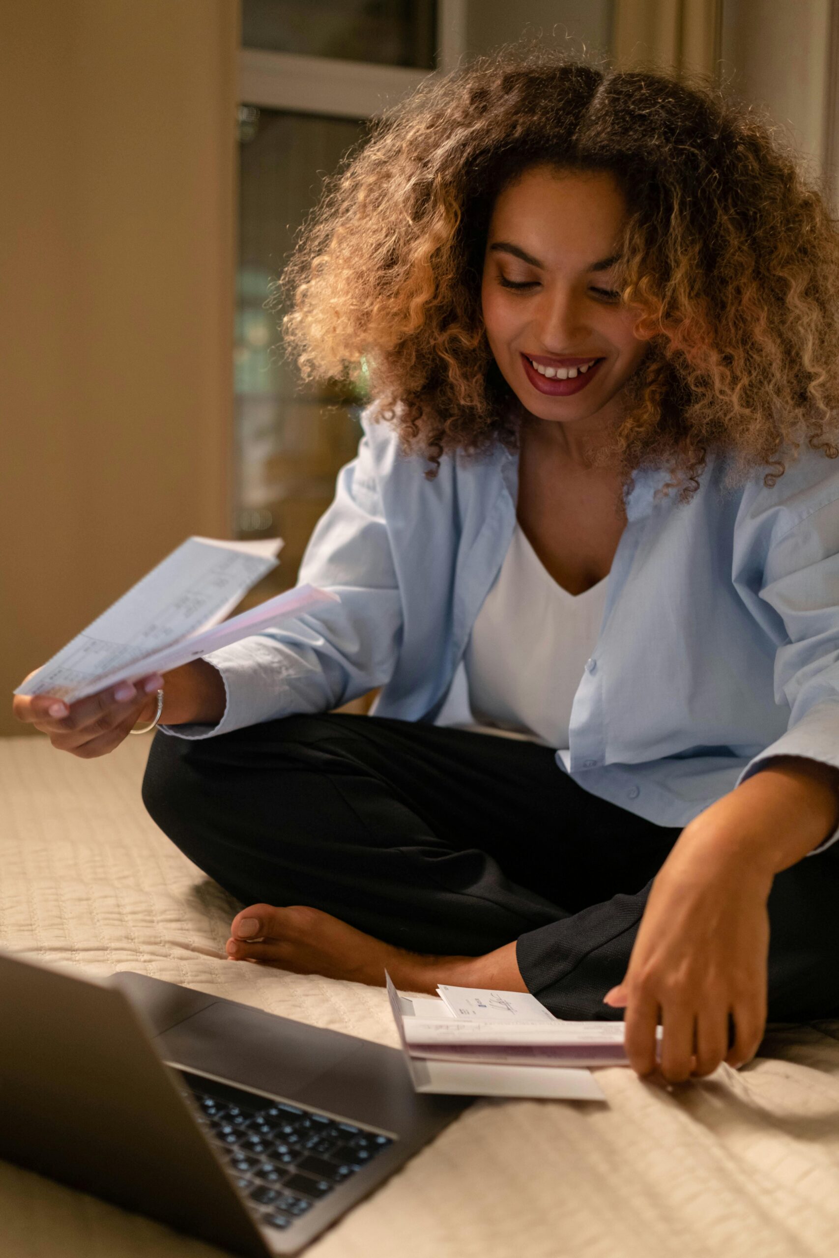 A cheerful woman with curly hair reads letters on her bed while using a laptop indoors.