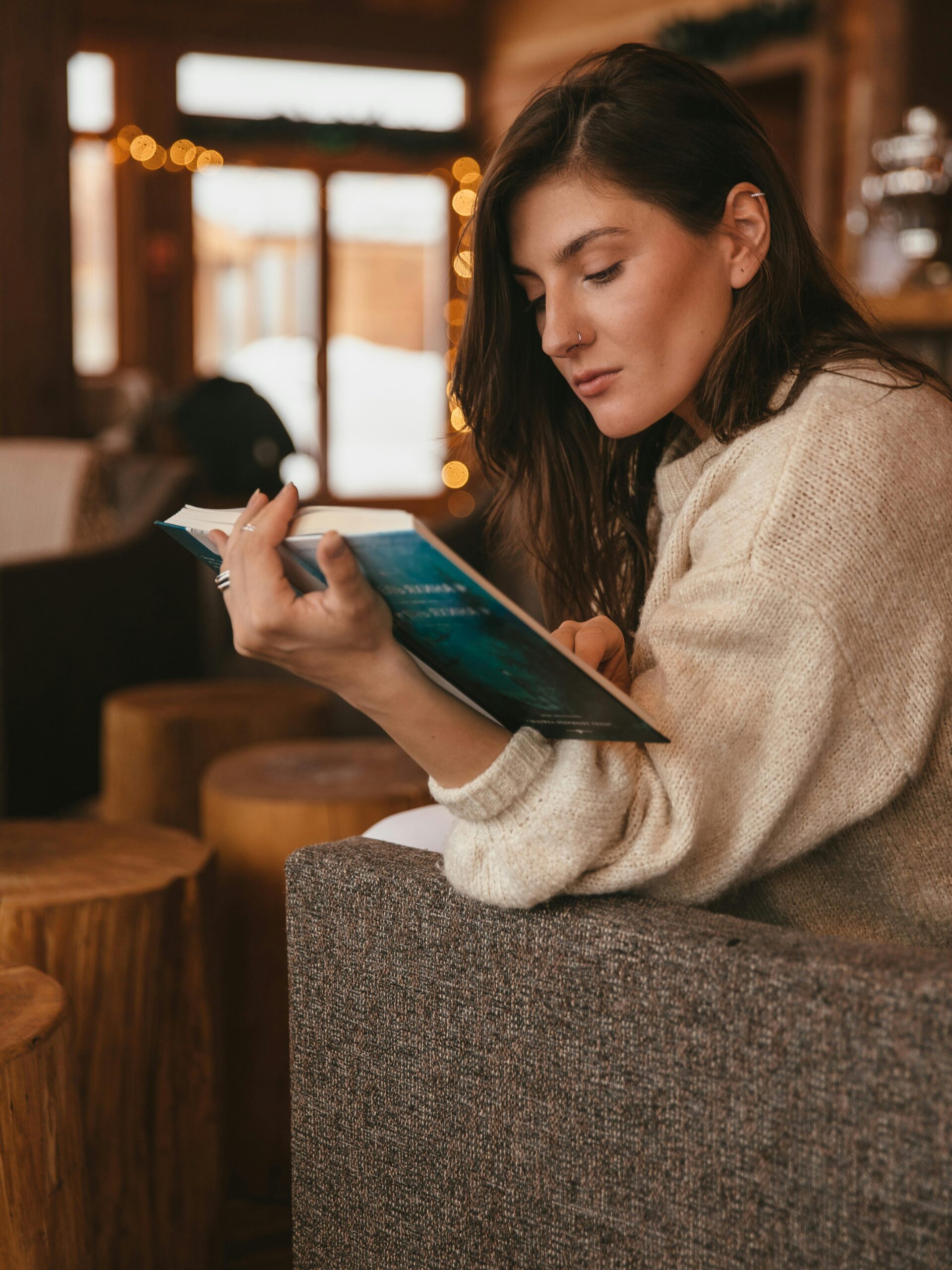 A woman in a cozy sweater reading a book in a warm indoor setting.
