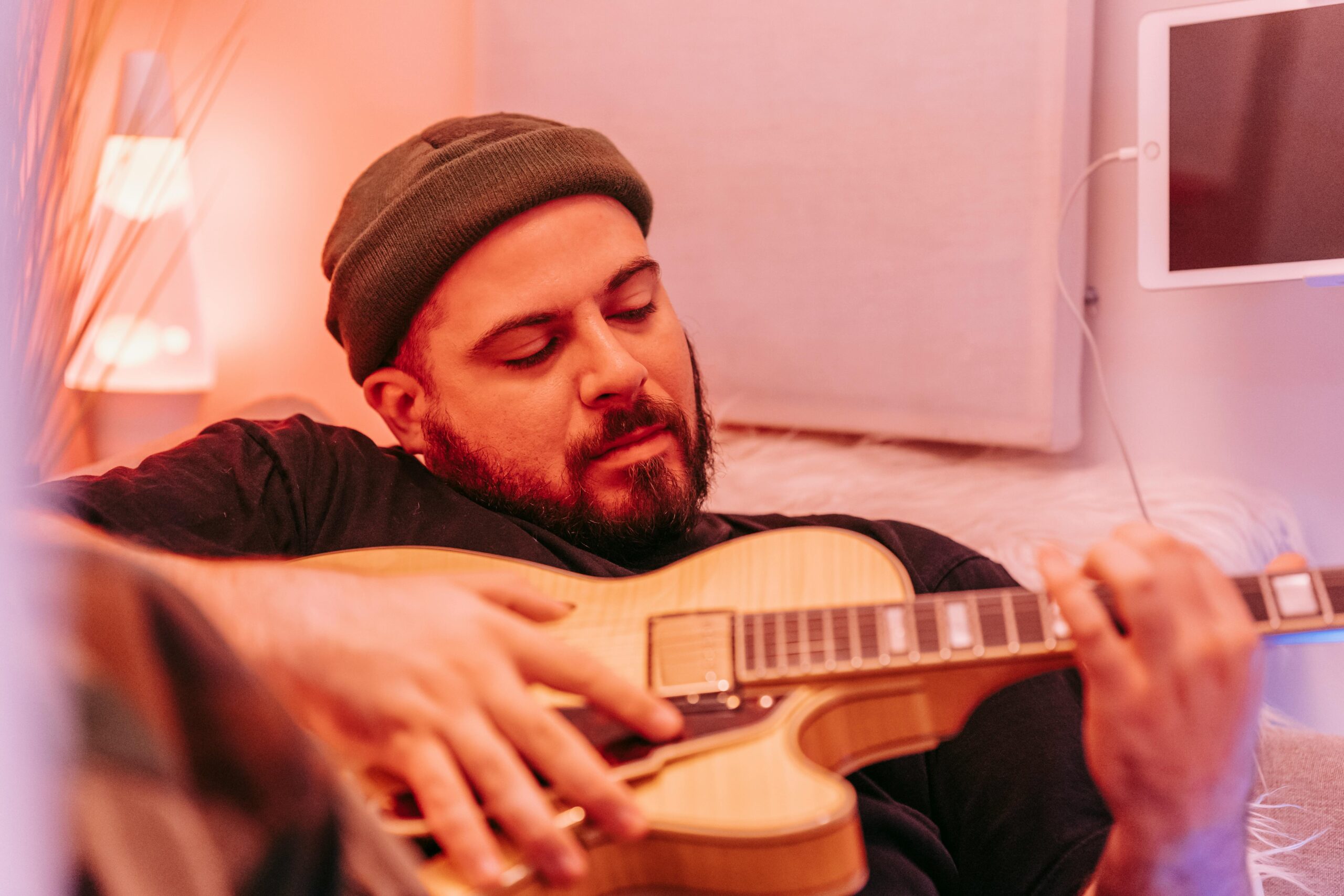 Man in beanie hat playing electric guitar, creating music in a cozy home studio.